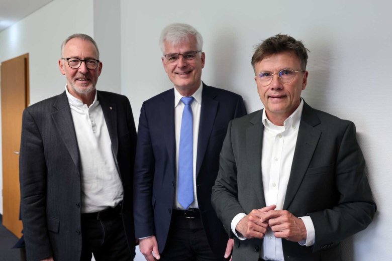 Drei Männer in Businesskleidung stehen nebeneinander in einem Innenraum und blicken in die Kamera. Alle tragen Sakkos und Hemden, zwei von ihnen tragen eine Brille. Der Hintergrund zeigt eine helle Wand und einen Flur, die Atmosphäre wirkt professionell und ruhig.