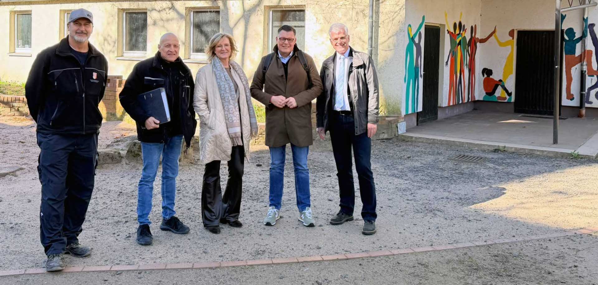 Fünf Erwachsene stehen nebeneinander auf einem Schulhof vor einem hellen Gebäude. Sie tragen Alltagskleidung wie Jacken und Mäntel und schauen in die Kamera. Rechts im Bild ist ein überdachter Eingangsbereich mit bunten Wandmalereien von stilisierten, tanzenden Figuren zu sehen. Die Szene ist sonnig, mit Schatten von Bäumen auf der Fassade.