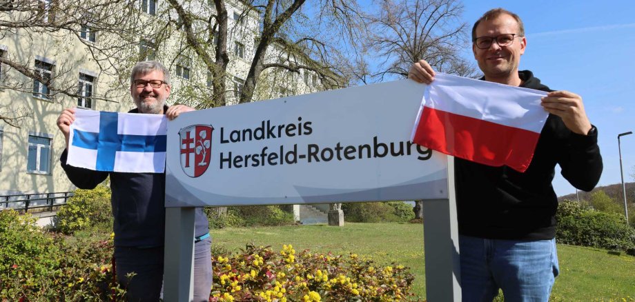 Zwei Männer stehen im Freien neben einem Schild mit der Aufschrift „Landkreis Hersfeld-Rotenburg“ und halten die Flaggen von Finnland und Polen; im Hintergrund sind ein Gebäude, Bäume und ein Garten zu sehen.