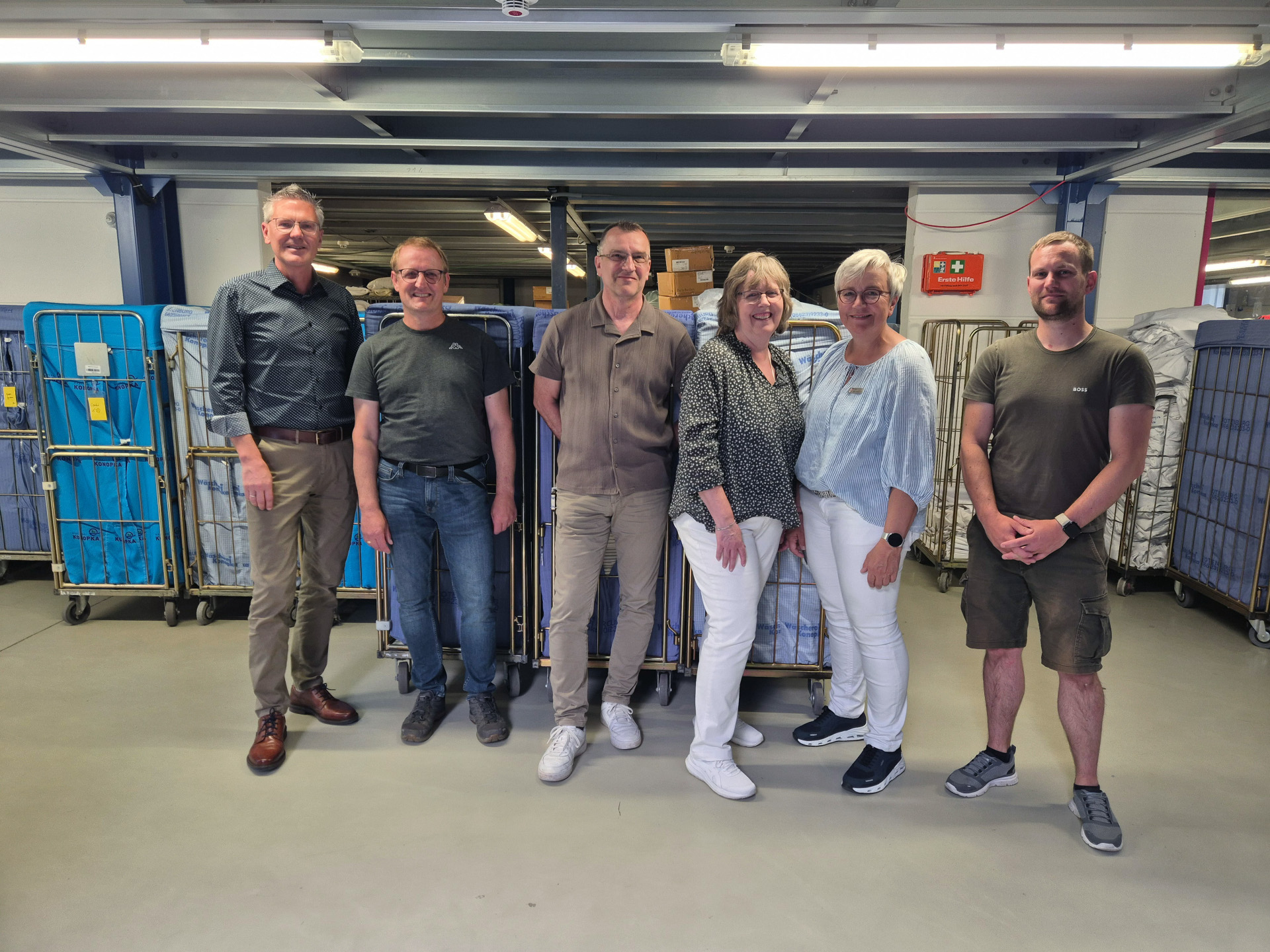 Sechs Personen stehen in einer Wäscherei vor mehreren großen Wäschecontainern. Hinter ihnen sind Arbeitsbereiche und Technik des Betriebs zu sehen. Von links stehen Erster Kreisbeigeordneter Dirk Noll, Seniorchef Markus Konopka, Andreas Olexiuk, Ulrike Hirschbrich, Hildegard Hehr-Willhardt und Juniorchef Jens Konopka.