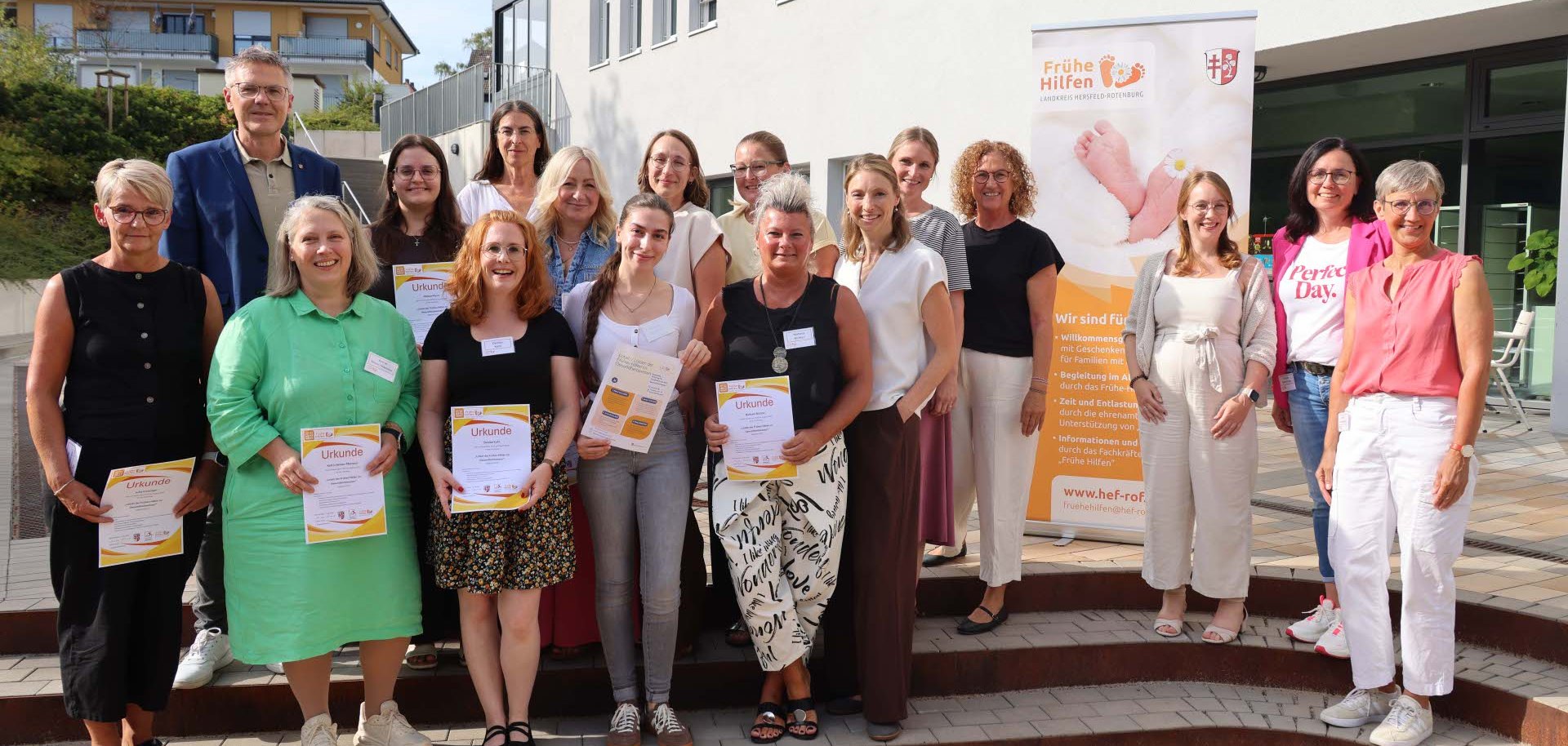 Rund 15 Personen, vor allem Frauen, stehen vor dem Landratsamt und halten Urkunden in der Hand. Es steht ein Rollup der Frühen Hilfen neben ihnen.