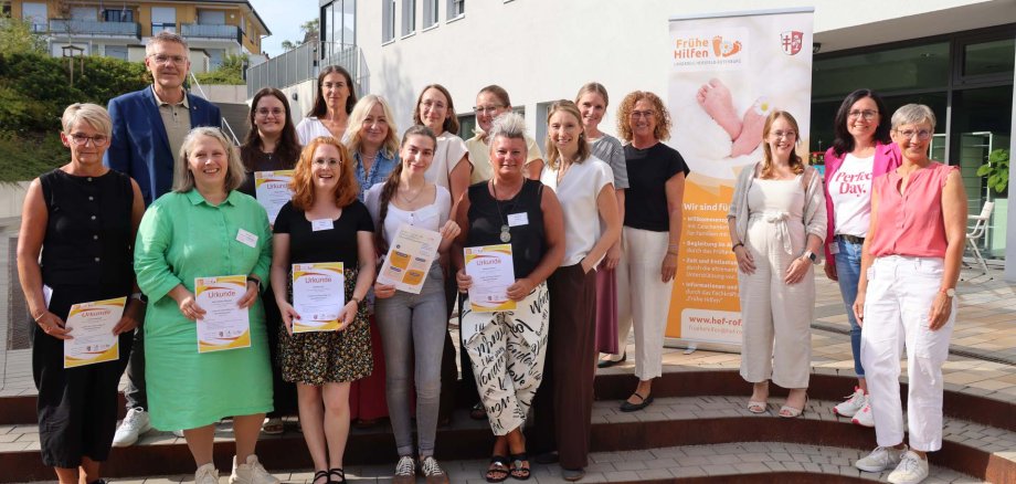 Rund 15 Personen, vor allem Frauen, stehen vor dem Landratsamt und halten Urkunden in der Hand. Es steht ein Rollup der Frühen Hilfen neben ihnen.