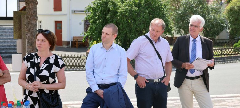 Vier Menschen, darunter eine Frau stehen, auf einem gepflasterten Hof. Im Bild sieht man einige Getränke-Flaschen und ein paar Gläser. Die Menschen blicken alle in Richtung eines Sprechenden, der auf dem Foto allerdings nicht zu sehen ist. Sie stehen neben einem Baum.