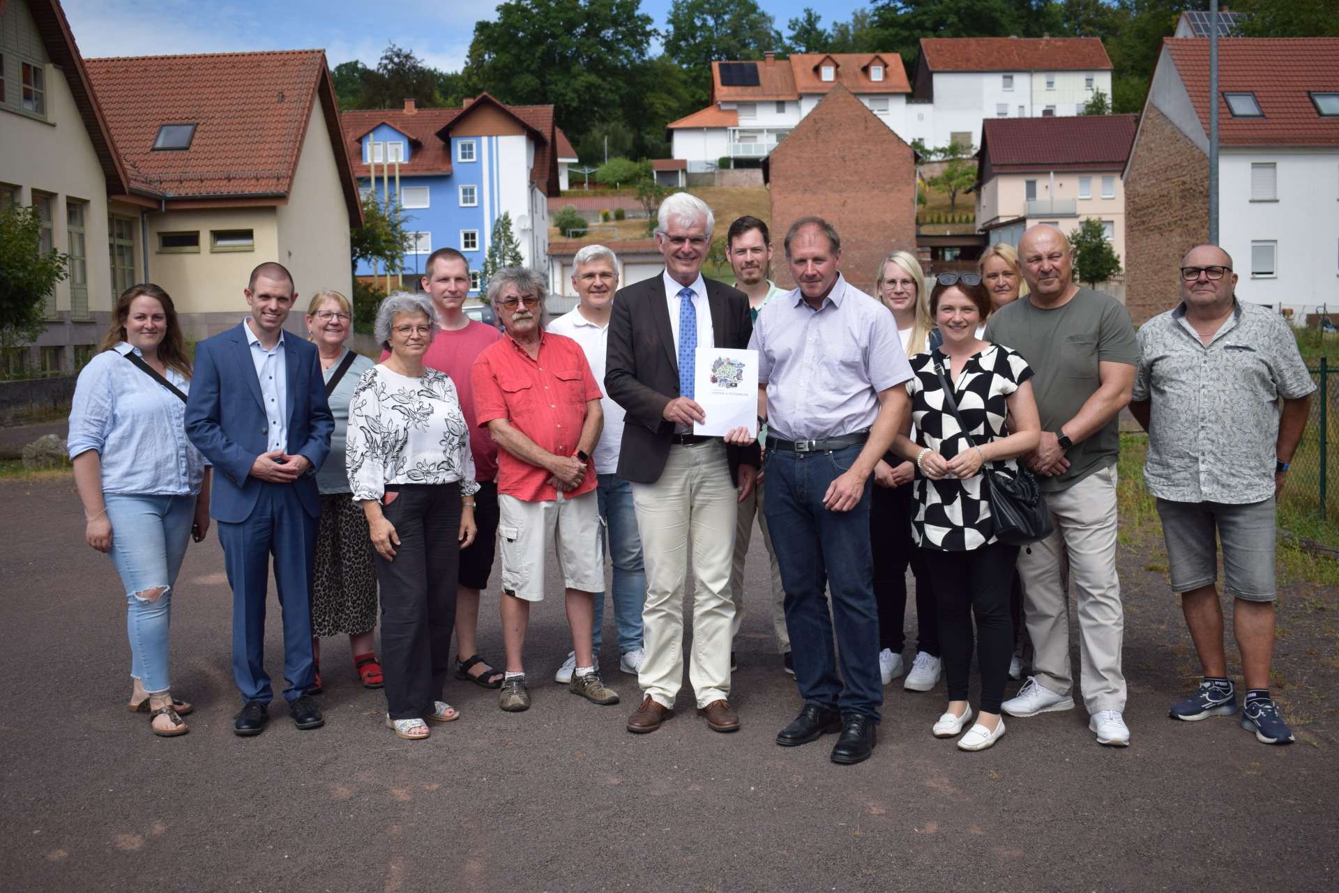 15 Menschen stehen auf einem Grundstück. Zwei Männer halten eine Mappe in der Hand. Im Hintergrund sieht man Häuser. 
