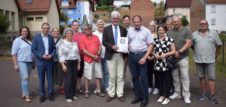 15 Menschen stehen auf einem Grundstück. Zwei Männer halten eine Mappe in der Hand. Im Hintergrund sieht man Häuser. 