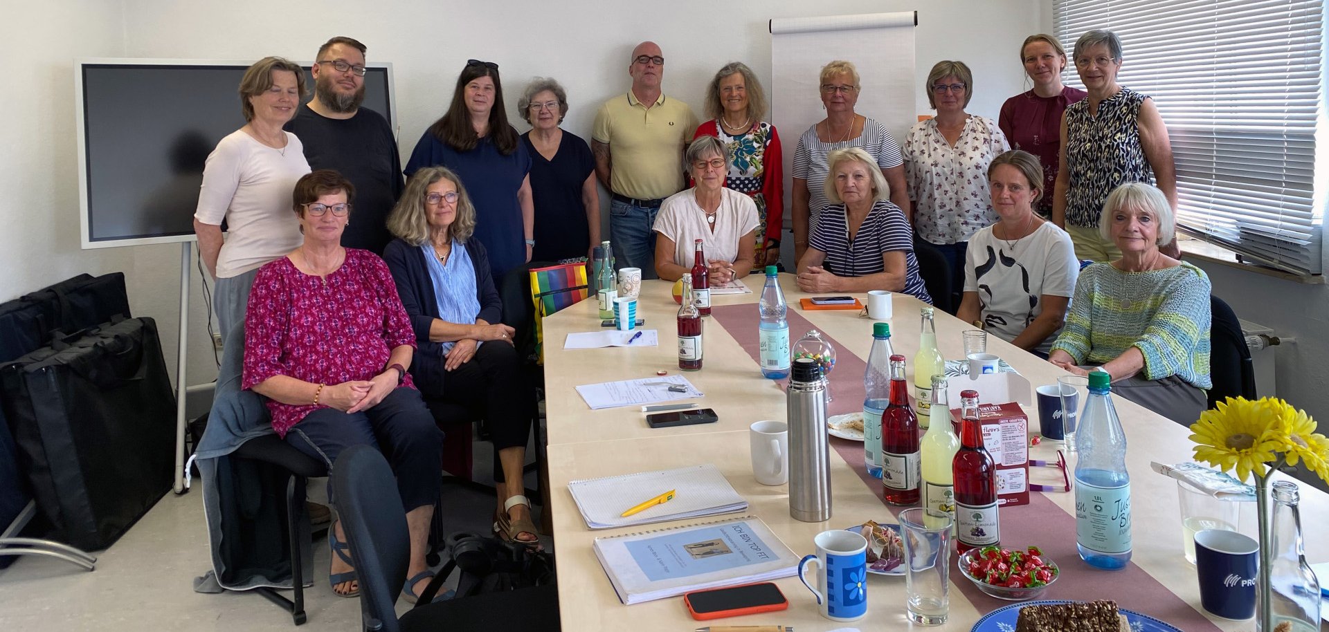 Gruppenfoto von etwa 15 Erwachsenen, die in einem hellen Besprechungsraum an mehreren zusammengestellten Tischen sitzen oder dahinter stehen. Auf den Tischen liegen Unterlagen, Getränke, Tassen und Snacks. Im Hintergrund sind ein Bildschirm, ein Flipchart und Fenster mit Jalousien zu sehen.
