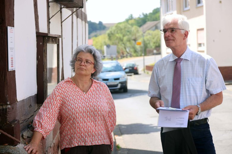 Ein Mann und eine Frau stehen in der Ortsmitte eines kleinen Ortes. Sie schauen jemandem beim Sprechen zu, der nicht im Bild zu sehen ist. Der Mann hält eine Mappe in der Hand.