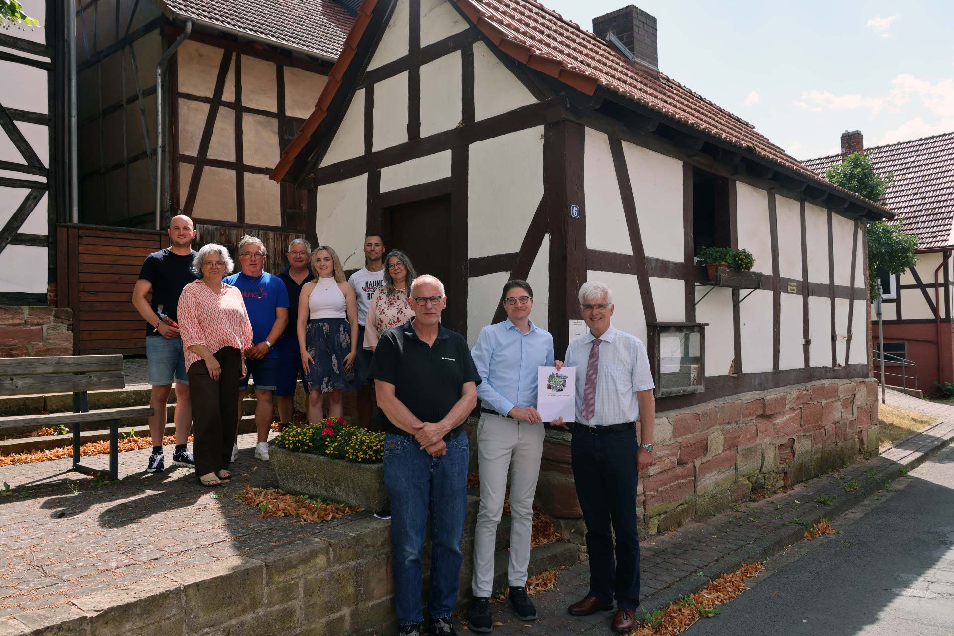 Mehrere Personen stehen vor einem alten Backhaus in der Ortsmitte eines Dorfes. Sie schauen in die Kamera und freuen sich.
