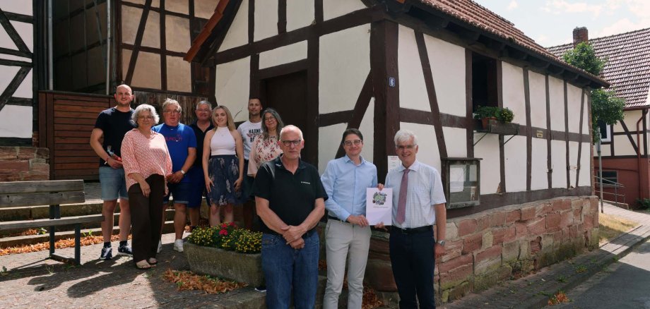Mehrere Personen stehen vor einem alten Backhaus in der Ortsmitte eines Dorfes. Sie schauen in die Kamera und freuen sich.
