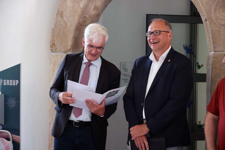 Zwei Männer stehen im Foyer eines Rathauses. Der eine Mann hält eine Mappe in der Hand aus der er etwas vorliest. Der andere Mann freut sich.