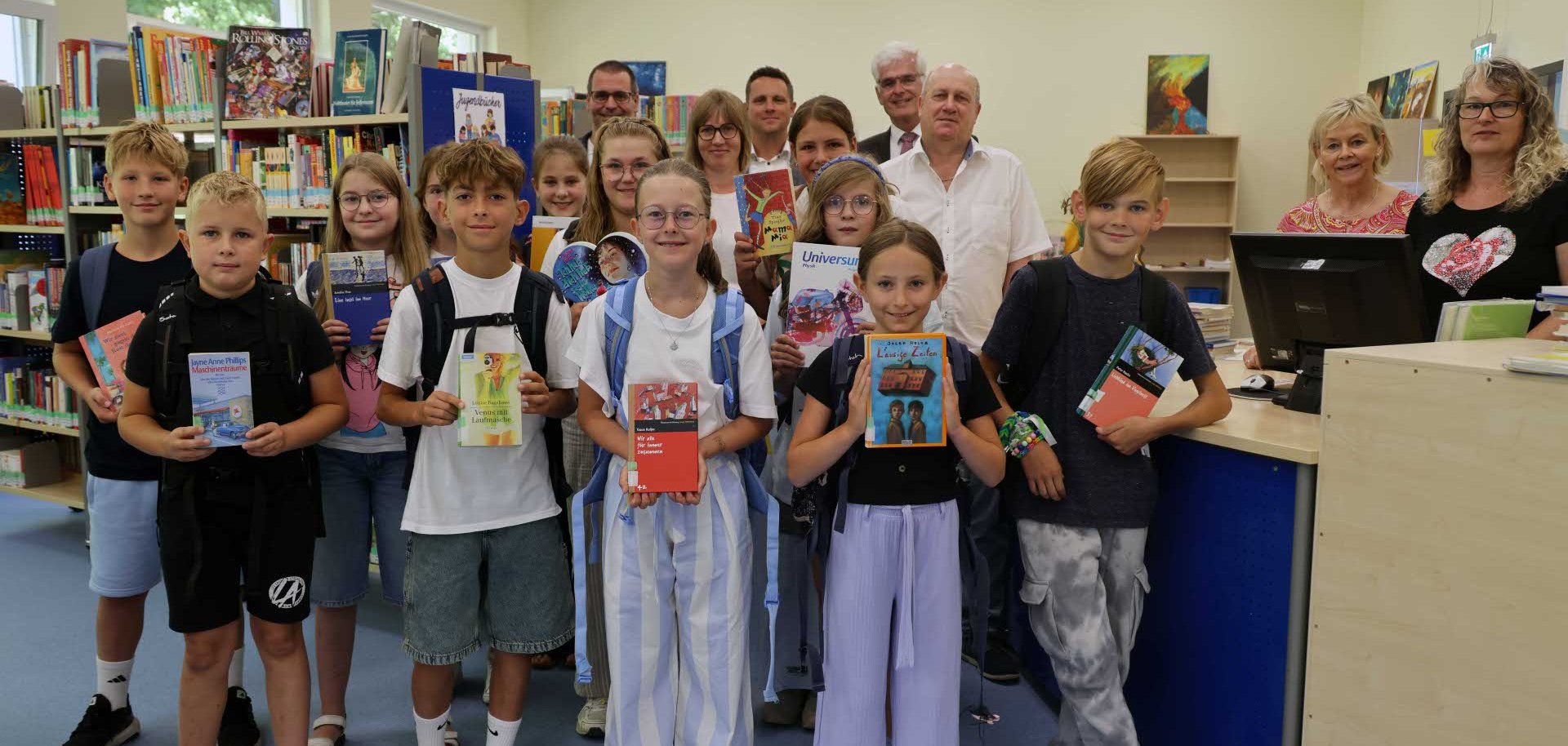 Gruppenfoto in einer Bibliothek: Mehrere Kinder und Jugendliche stehen gemeinsam mit Erwachsenen zwischen Bücherregalen und vor einem Empfangstresen. Einige der Kinder halten Bücher in die Kamera. Im Hintergrund sind weitere Bücherregale, Sitz- und Arbeitsbereiche sowie Kunstwerke an den Wänden zu sehen.