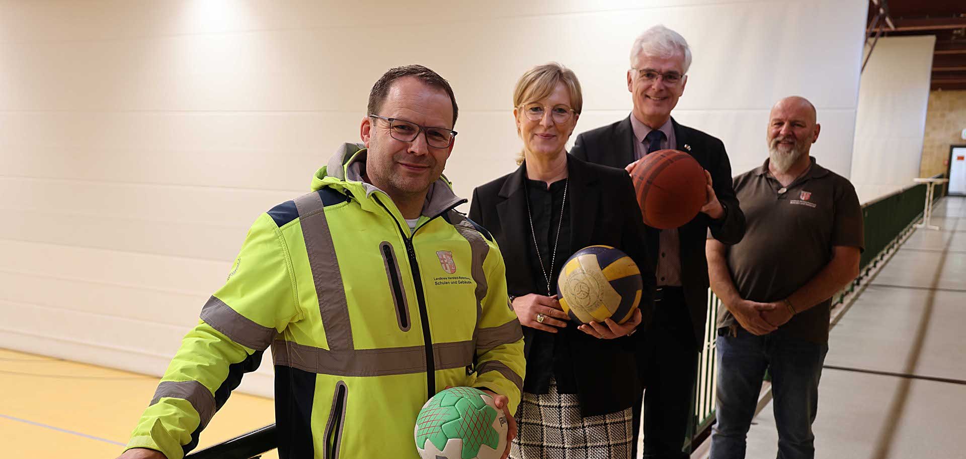Vier Personen stehen in einer Sporthalle. Drei von ihnen halten jeweils einen Ball. Im Hintergrund sieht man eine Sporthalle.
