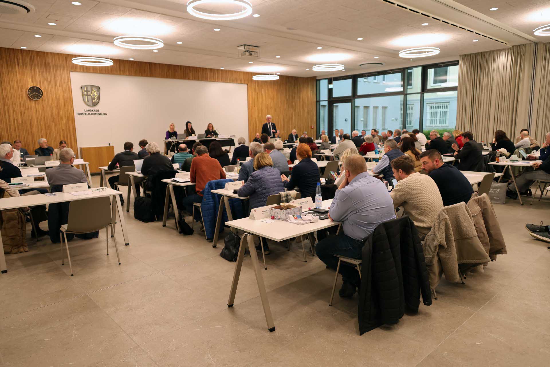 Großer Sitzungssaal mit zahlreichen Personen an Tischen, die an einer Kreistagssitzung teilnehmen. Im Vordergrund sitzen Zuhörerinnen und Zuhörer, im Hintergrund spricht ein Redner am Pult vor dem Präsidium.