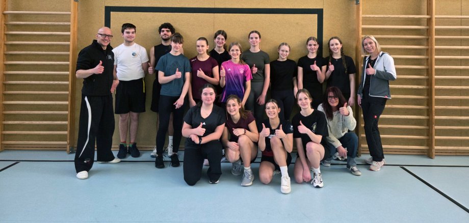 Eine Gruppe von Jugendlichen und zwei Erwachsenen posiert in einer Sporthalle unter einem Basketballkorb. Einige stehen, andere knien vorne, und fast alle zeigen mit einem Lächeln den Daumen nach oben. Im Hintergrund sind Sprossenwände und die Hallenwand zu sehen.