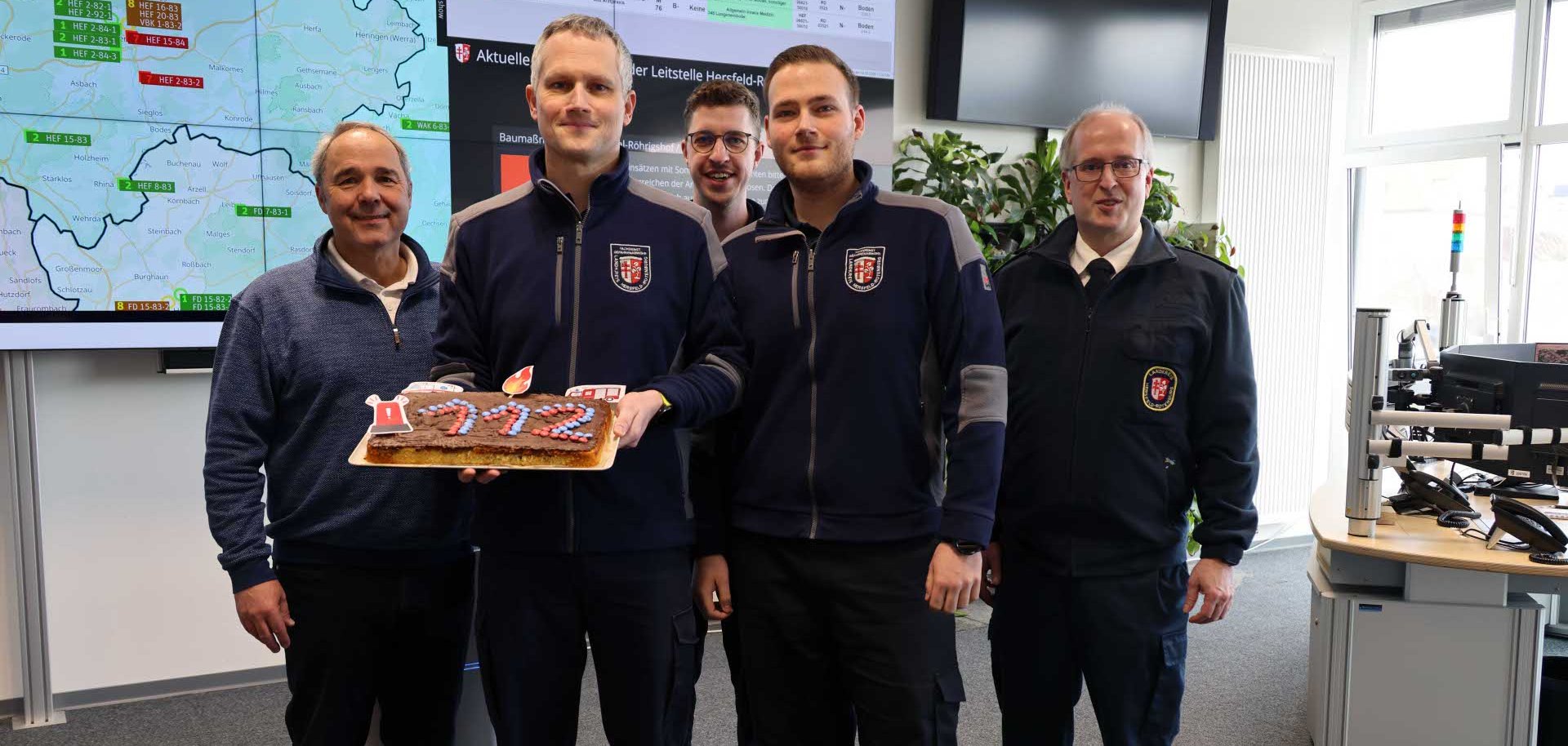 Fünf Männer in Einsatzkleidung stehen in einer Leitstelle vor großen Monitoren mit Karten und Einsatzdaten. Zwei von ihnen halten einen Kuchen mit der Zahl ‚112‘, dem Symbol des Notrufs. Die Gruppe lächelt in die Kamera.