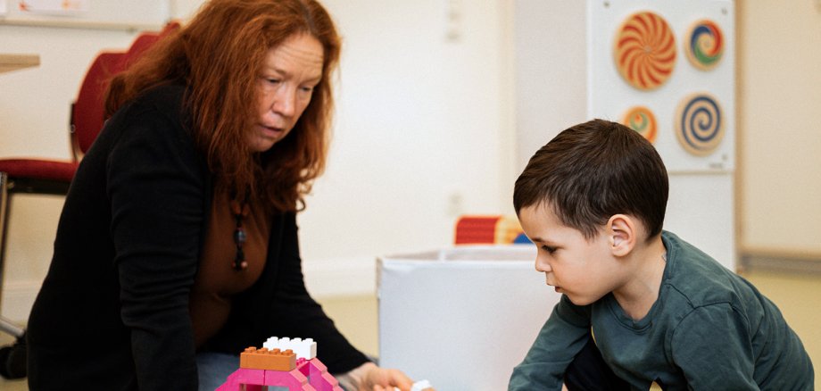 Zwei Personen sind auf dem Bild zu erkennen, die miteinander spielen. Es ist zudem Spielzeug im Vordergrund zu erkennen. Es ist eine erwachsene Person und ein Kind zu sehen.