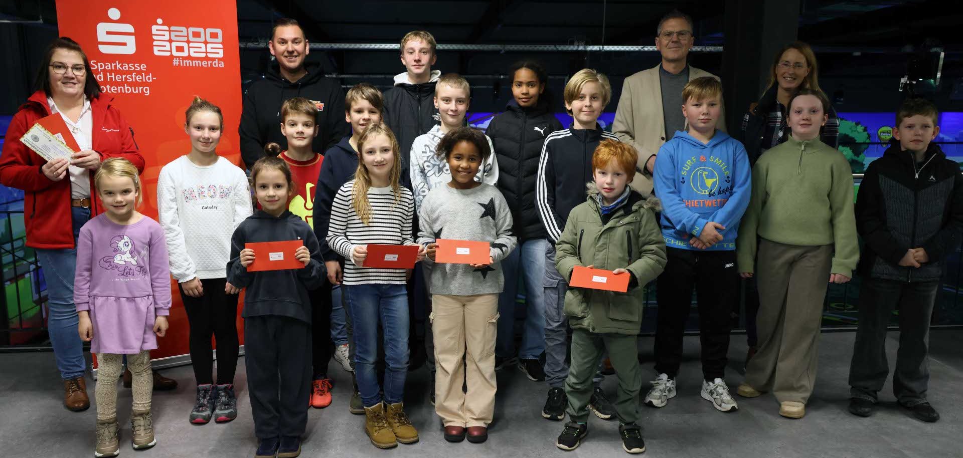 Eine Gruppe von Kindern und Jugendlichen steht gemeinsam mit einigen Erwachsenen in einer Indoor-Kartbahn. Einige Kinder halten rote Umschläge mit Gewinnen in den Händen. Im Hintergrund ist ein Banner der Sparkasse Hersfeld-Rotenburg zu sehen. Die Gruppe posiert lächelnd für ein gemeinsames Erinnerungsfoto.