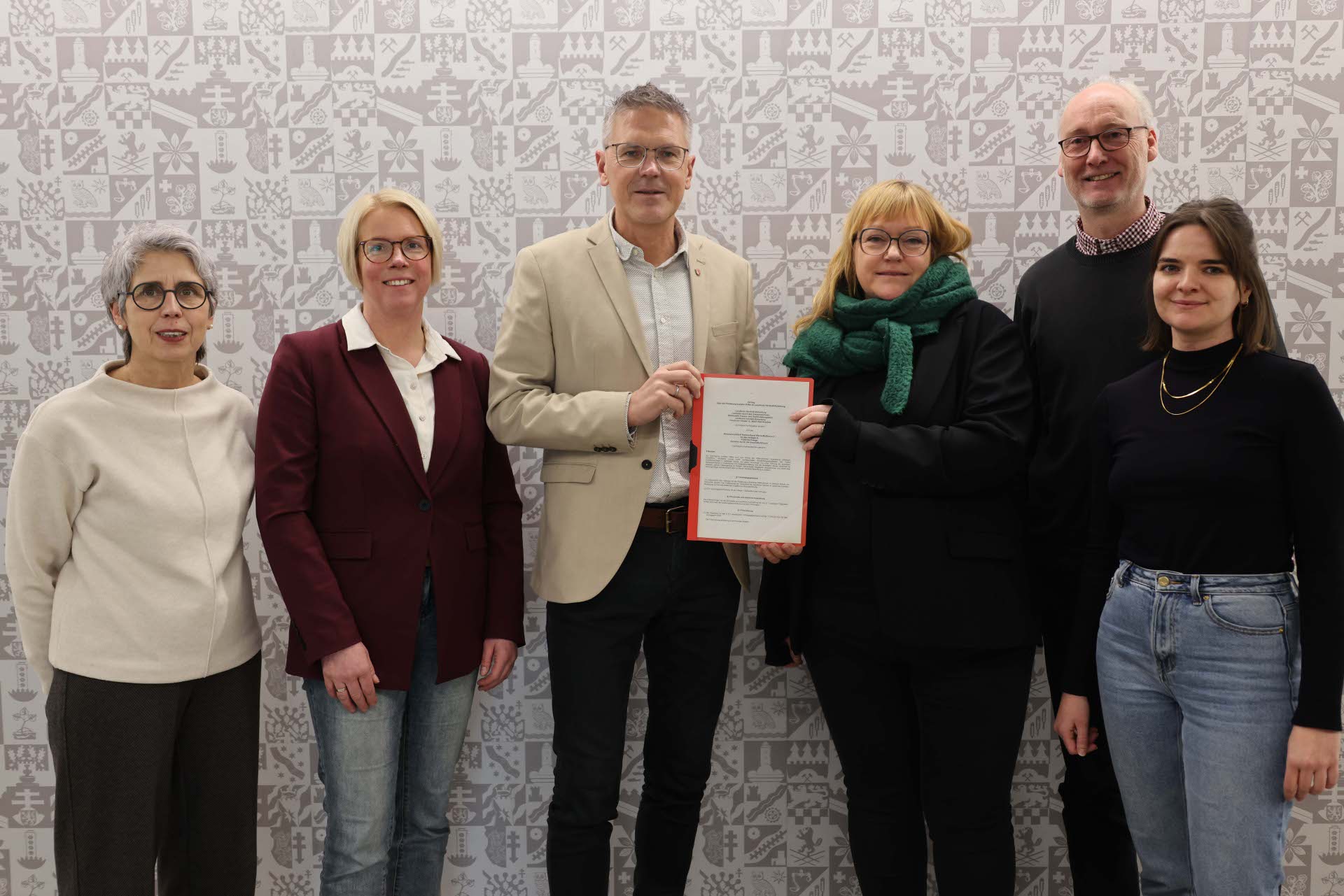 Sechs Personen stehen nebeneinander vor einer hell gemusterten Wand. In der Mitte hält ein Mann gemeinsam mit einer Frau ein Dokument in den Händen. Die Gruppe blickt in die Kamera und wirkt sachlich und freundlich. Die Personen tragen überwiegend schlichte, elegante Alltagskleidung.