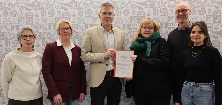 Sechs Personen stehen nebeneinander vor einer hell gemusterten Wand. In der Mitte hält ein Mann gemeinsam mit einer Frau ein Dokument in den Händen. Die Gruppe blickt in die Kamera und wirkt sachlich und freundlich. Die Personen tragen überwiegend schlichte, elegante Alltagskleidung.