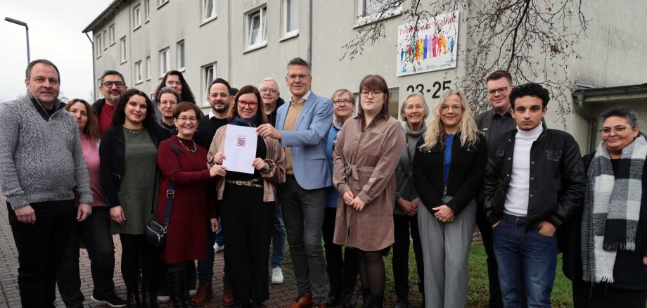 Eine Gruppe Menschen steht vor einem Gebäude. Die meisten Personen lächeln und es wird ein Schriftstück feierlich übergeben.