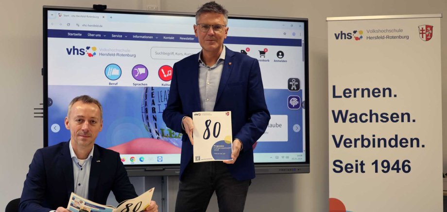 Auf dem Foto sind zwei Männer zu sehen. Beide haben eine Ausgabe des vhs-Programms in ihrer Hand. Ein Mann sitzt und einer steht. Sie sind in einem Kursraum, im Hintergrund ein großer Bildschirm und im rechten Teil des Bildes ein Roll-Up.