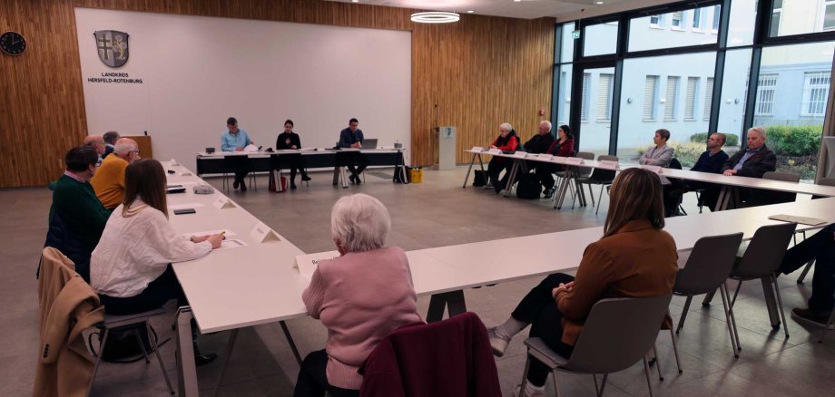 In einem Raum sitzen verschiedene Personen in einem Besprechungsraum. Im Hintergrund ist eine Fensterfront zu sehen, sowie eine braune Wand.