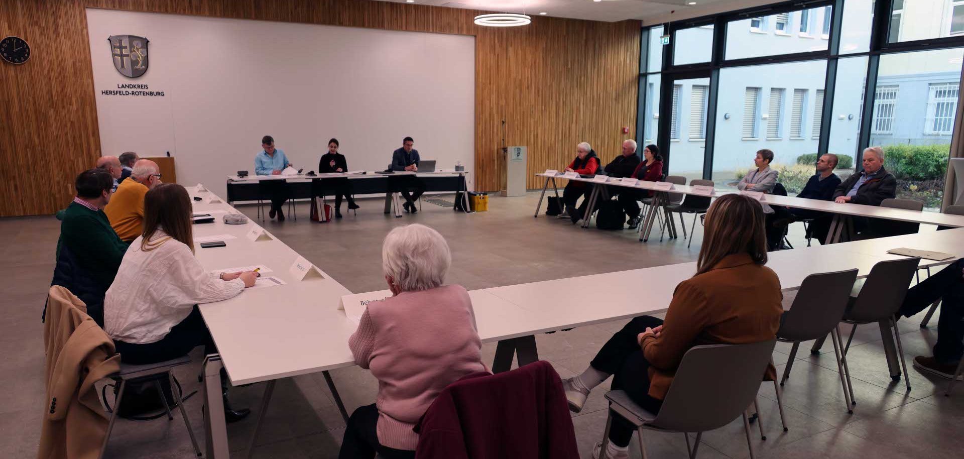 In einem Raum sitzen verschiedene Personen in einem Besprechungsraum. Im Hintergrund ist eine Fensterfront zu sehen, sowie eine braune Wand.