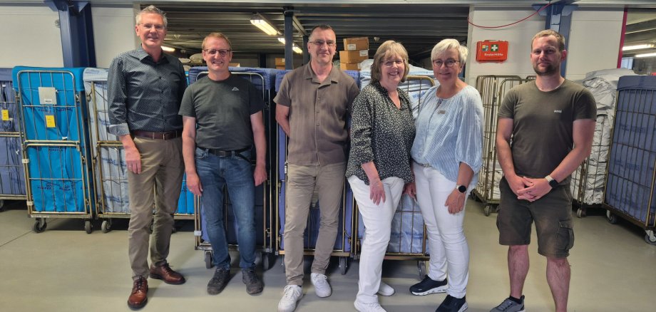 Sechs Personen stehen in einer Wäscherei vor mehreren großen Wäschecontainern. Hinter ihnen sind Arbeitsbereiche und Technik des Betriebs zu sehen. Von links stehen Erster Kreisbeigeordneter Dirk Noll, Seniorchef Markus Konopka, Andreas Olexiuk, Ulrike Hirschbrich, Hildegard Hehr-Willhardt und Juniorchef Jens Konopka.