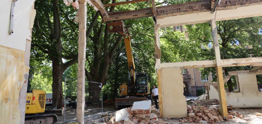 Ein Bagger setzt an, weitere Teile einer alten Turnhalle abzureißen. Im Bild sieht man viel Bauschutt, einen Bagger und einen Menschen, der sich den Vorgang anschaut.