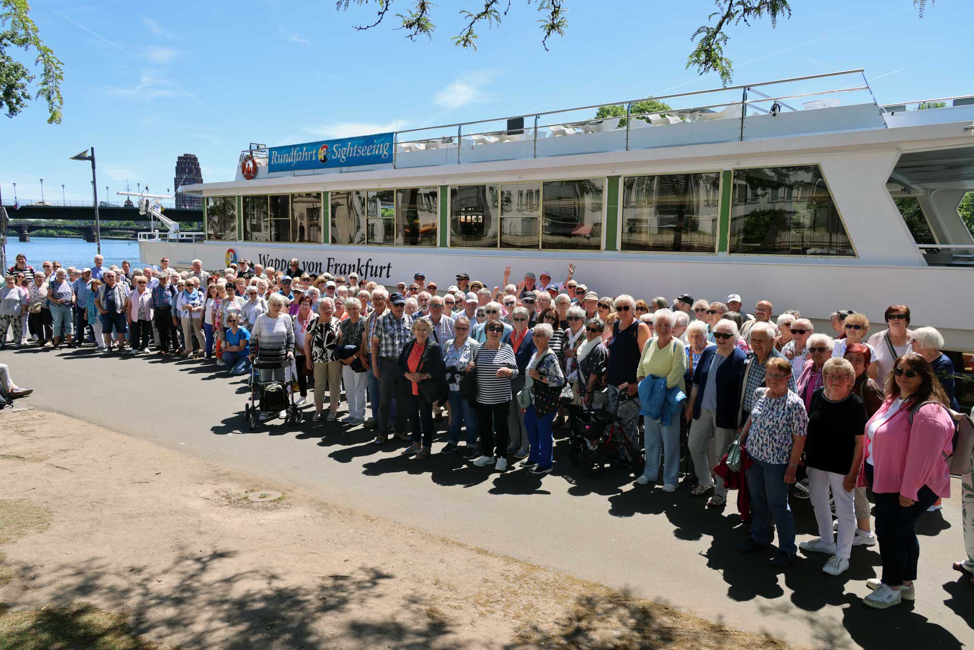 Eine große Reisegruppe steht am Ufer vor einem weißen Ausflugsschiff mit der Aufschrift „Wappen von Frankfurt“ und blickt in die Kamera. Im Hintergrund sind der Fluss und eine Brücke zu sehen.