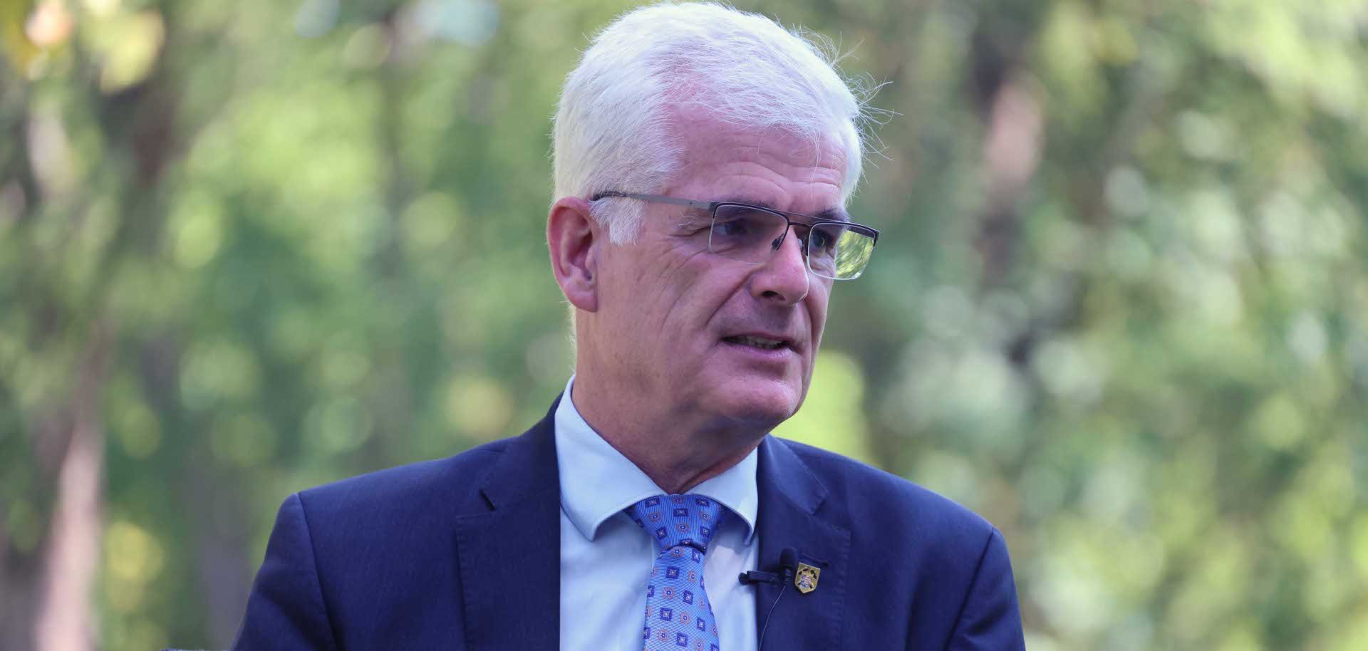 Ein älterer Mann mit kurzen weißen Haaren und randloser Brille sitzt im Freien auf einem Stuhl. Er trägt einen dunkelblauen Anzug, ein hellblaues Hemd und eine hellblaue Krawatte mit dezentem Muster. An seinem Jackett sind ein kleines Ansteckabzeichen und ein Ansteckmikrofon befestigt. Er blickt leicht zur Seite und scheint zu sprechen. Der unscharfe Hintergrund zeigt grüne Bäume und vermittelt den Eindruck eines Interviews oder Gesprächs in einer natürlichen Umgebung.
