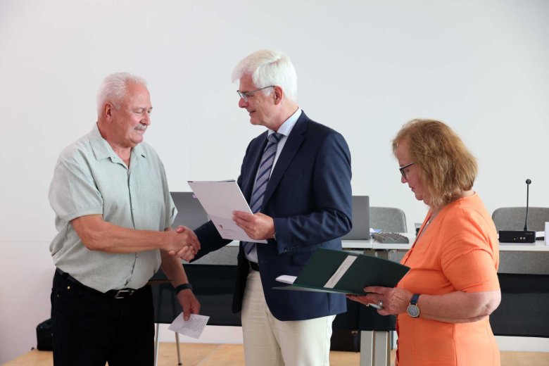 Ein Mann gibt einem anderen Mann die Hand und hat dabei eine Mappe in der Hand. Eine Frau, in einem orangenen Kleid, schaut auf Unterlagen in ihrer Hand.