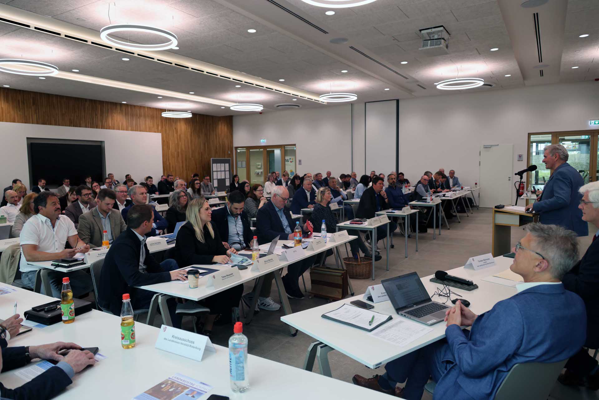 Ein großer Konferenzraum mit mehreren Reihen von Tischen, an denen zahlreiche Personen in formeller Kleidung sitzen und einer Präsentation zuhören. Im Vordergrund sitzt ein Mann im blauen Anzug an einem Tisch mit Laptop und Unterlagen. Rechts spricht ein Redner an einem Mikrofon zu den Teilnehmenden. Die moderne Veranstaltung findet in einem hellen Raum mit Holzverkleidung, großen Deckenleuchten und professioneller Tagungsausstattung statt.