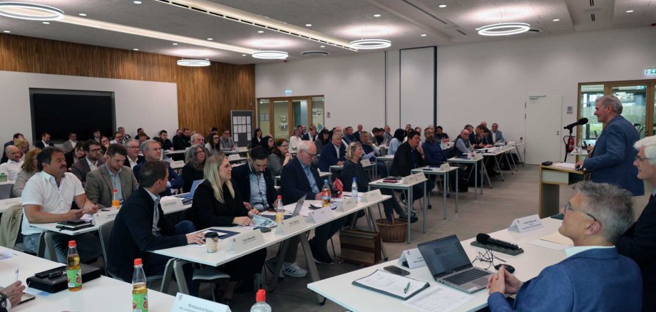 Ein großer Konferenzraum mit mehreren Reihen von Tischen, an denen zahlreiche Personen in formeller Kleidung sitzen und einer Präsentation zuhören. Im Vordergrund sitzt ein Mann im blauen Anzug an einem Tisch mit Laptop und Unterlagen. Rechts spricht ein Redner an einem Mikrofon zu den Teilnehmenden. Die moderne Veranstaltung findet in einem hellen Raum mit Holzverkleidung, großen Deckenleuchten und professioneller Tagungsausstattung statt.