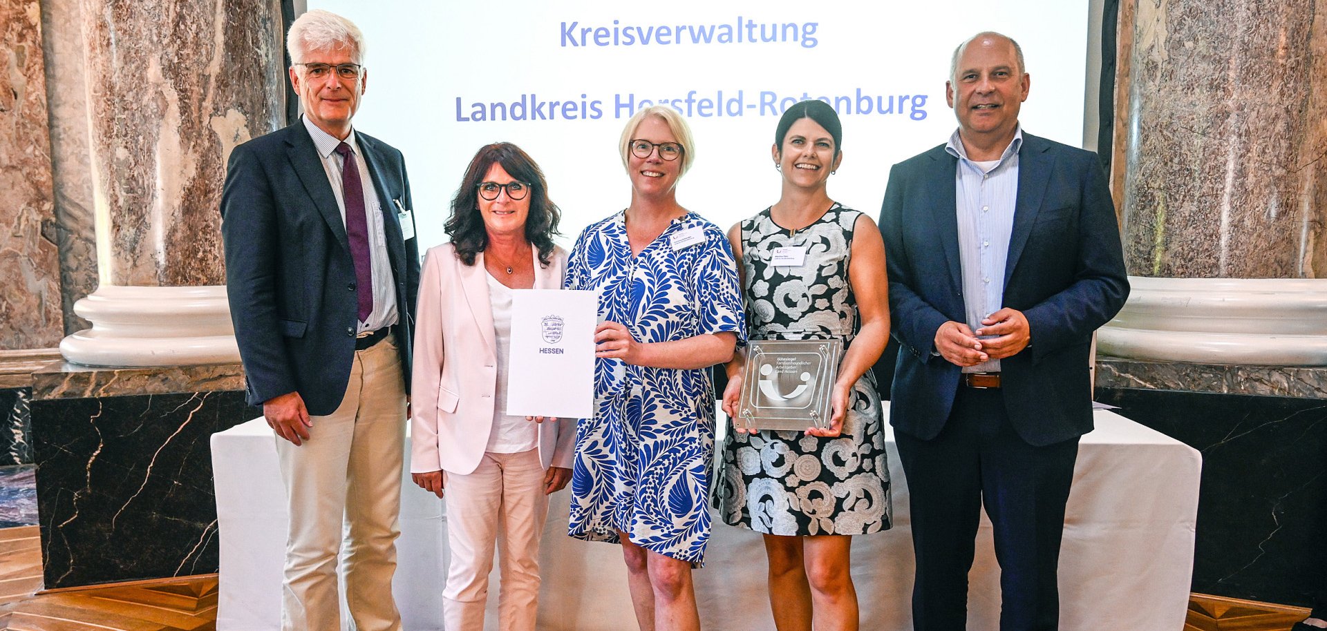 Fünf Personen stehen bei einer Preisverleihung vor einer Leinwand mit der Aufschrift „Kreisverwaltung Landkreis Hersfeld-Rotenburg“. Zwei Frauen in der Mitte halten eine Urkunde des Landes Hessen sowie eine gläserne Auszeichnung in die Kamera. Die drei weiteren Personen stehen daneben und lächeln. Die Aufnahme entstand in einem repräsentativen Saal mit Marmorsäulen und Parkettboden.