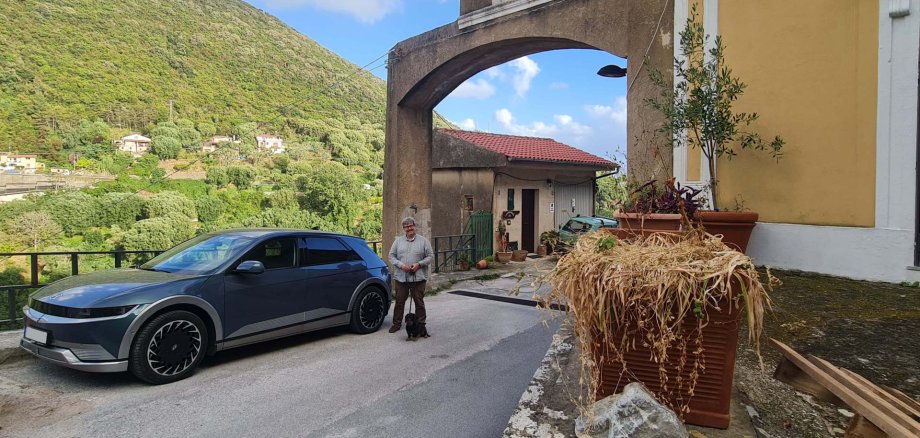 Eine Person steht mit einem kleinen Hund neben einem geparkten Elektroauto auf einer schmalen Straße. Im Hintergrund sind eine Kirche mit Glockenturm, Häuser und bewaldete Hügel zu sehen. Die Szene vermittelt den Eindruck einer Reise durch eine ländliche Bergregion.