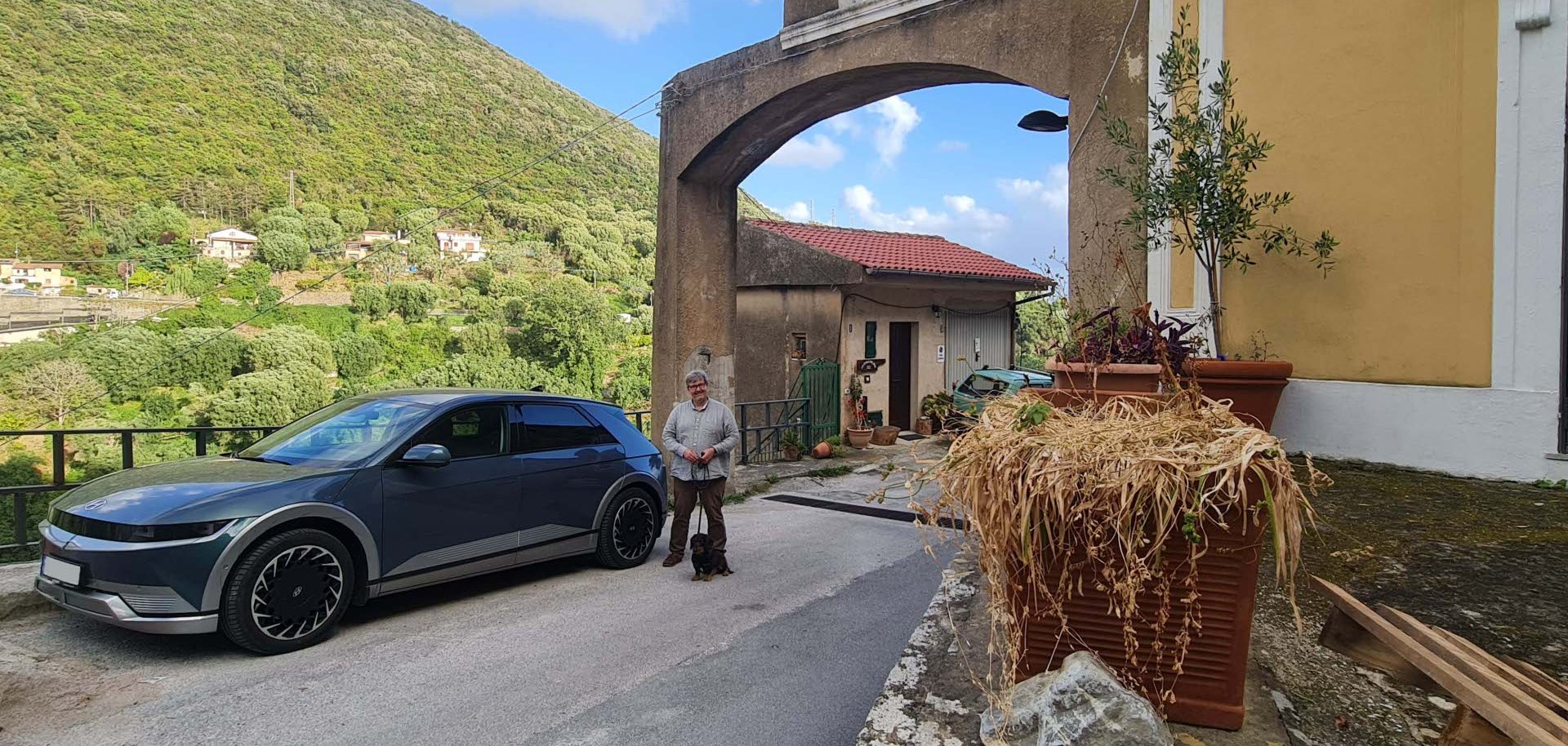 Eine Person steht mit einem kleinen Hund neben einem geparkten Elektroauto auf einer schmalen Straße. Im Hintergrund sind eine Kirche mit Glockenturm, Häuser und bewaldete Hügel zu sehen. Die Szene vermittelt den Eindruck einer Reise durch eine ländliche Bergregion.
