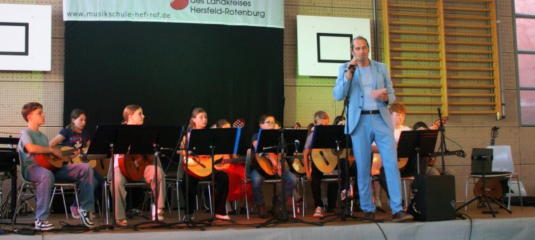 Auf einer Bühne sitzen mehrere Kinder mit Gitarren hinter Notenständern und bereiten einen Auftritt vor. Rechts steht ein Mann in einem hellblauen Anzug mit Mikrofon und Notizzettel. Im Hintergrund hängt ein Banner der Musikschule des Landkreises Hersfeld-Rotenburg in einer Turnhalle.
