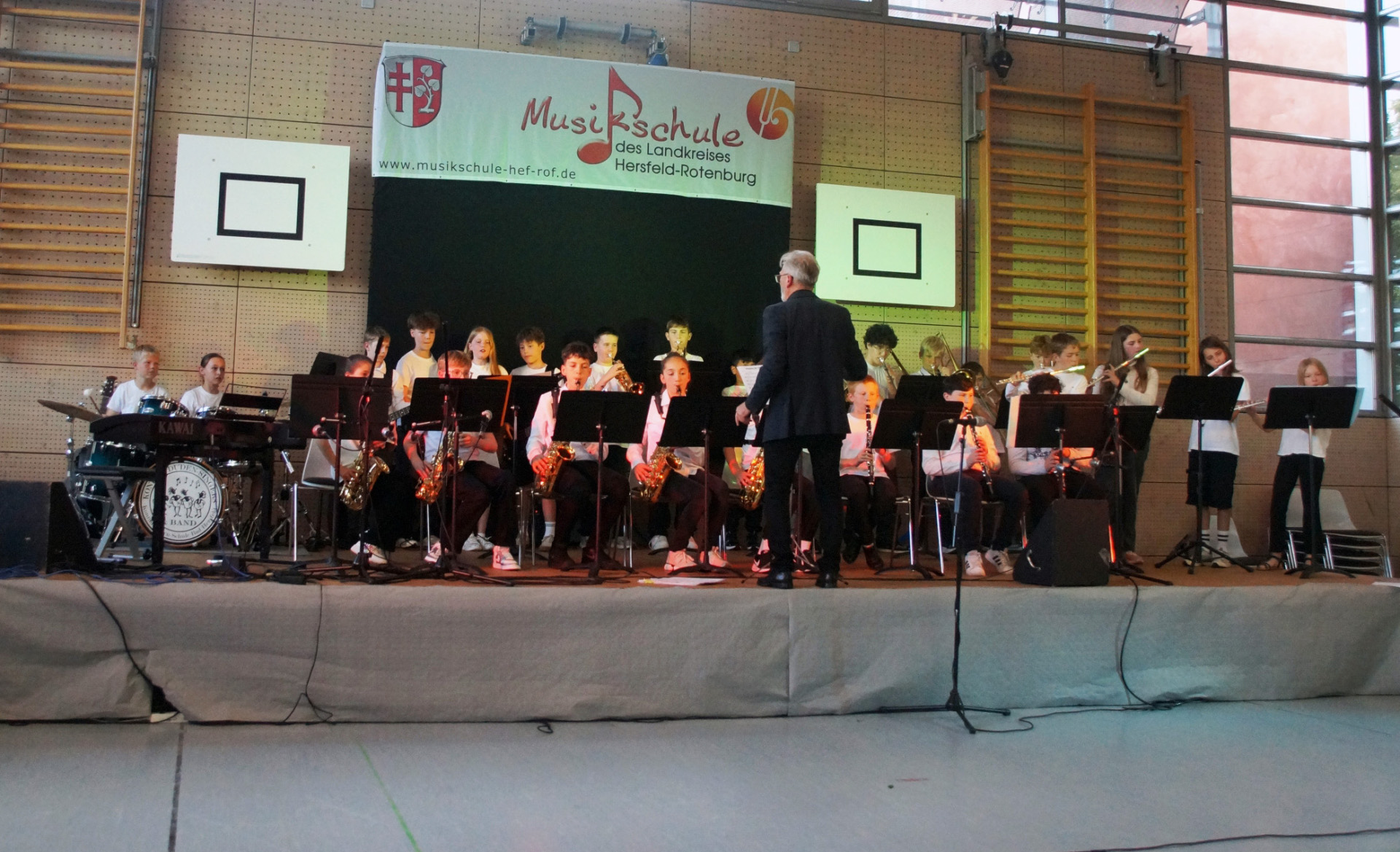 Auf einer Bühne in einer Turnhalle musizieren Kinder und Jugendliche in einem Bläserensemble mit Saxophonen, Klarinetten, Flöten und Schlagzeug hinter Notenständern. Ein Dirigent steht mit dem Rücken zum Publikum vor dem Ensemble. Im Hintergrund hängt ein Banner der Musikschule des Landkreises Hersfeld-Rotenburg.