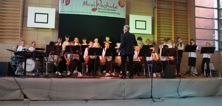 Auf einer Bühne in einer Turnhalle musizieren Kinder und Jugendliche in einem Bläserensemble mit Saxophonen, Klarinetten, Flöten und Schlagzeug hinter Notenständern. Ein Dirigent steht mit dem Rücken zum Publikum vor dem Ensemble. Im Hintergrund hängt ein Banner der Musikschule des Landkreises Hersfeld-Rotenburg.