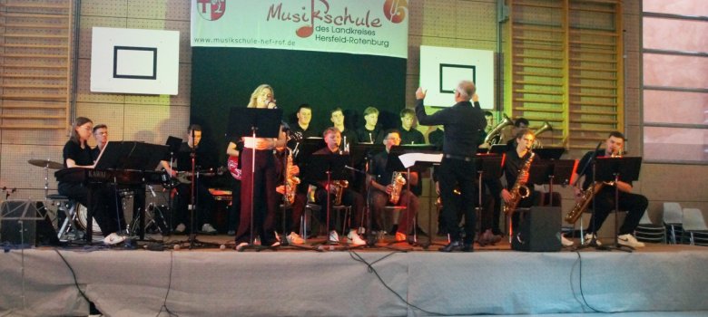 Auf einer Bühne in einer Turnhalle tritt eine Jugend-Jazzband auf. Eine Sängerin steht am Mikrofon, während ein Dirigent mit erhobenen Armen das Ensemble leitet. Die Musikerinnen und Musiker spielen unter anderem Klavier, Schlagzeug, E-Gitarre, Saxophone und Blechblasinstrumente. Im Hintergrund hängt ein Banner der Musikschule des Landkreises Hersfeld-Rotenburg.