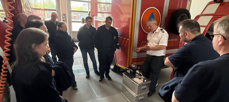 Eine Gruppe von Feuerwehr- und Katastrophenschutzkräften steht in einer Fahrzeughalle der Feuerwehr und verfolgt aufmerksam die Erläuterungen eines Ausbilders. Dieser präsentiert vor einem rot-weißen Fahrzeug des Zivilschutzes verschiedene Messgeräte und Ausrüstungskoffer. Die Teilnehmenden stehen im Halbkreis um die Vorführung und erhalten Einblicke in spezielle Messtechnik für Gefahrstofflagen. Im Hintergrund sind weitere Einsatzfahrzeuge und die geöffnete Fahrzeughalle zu sehen.