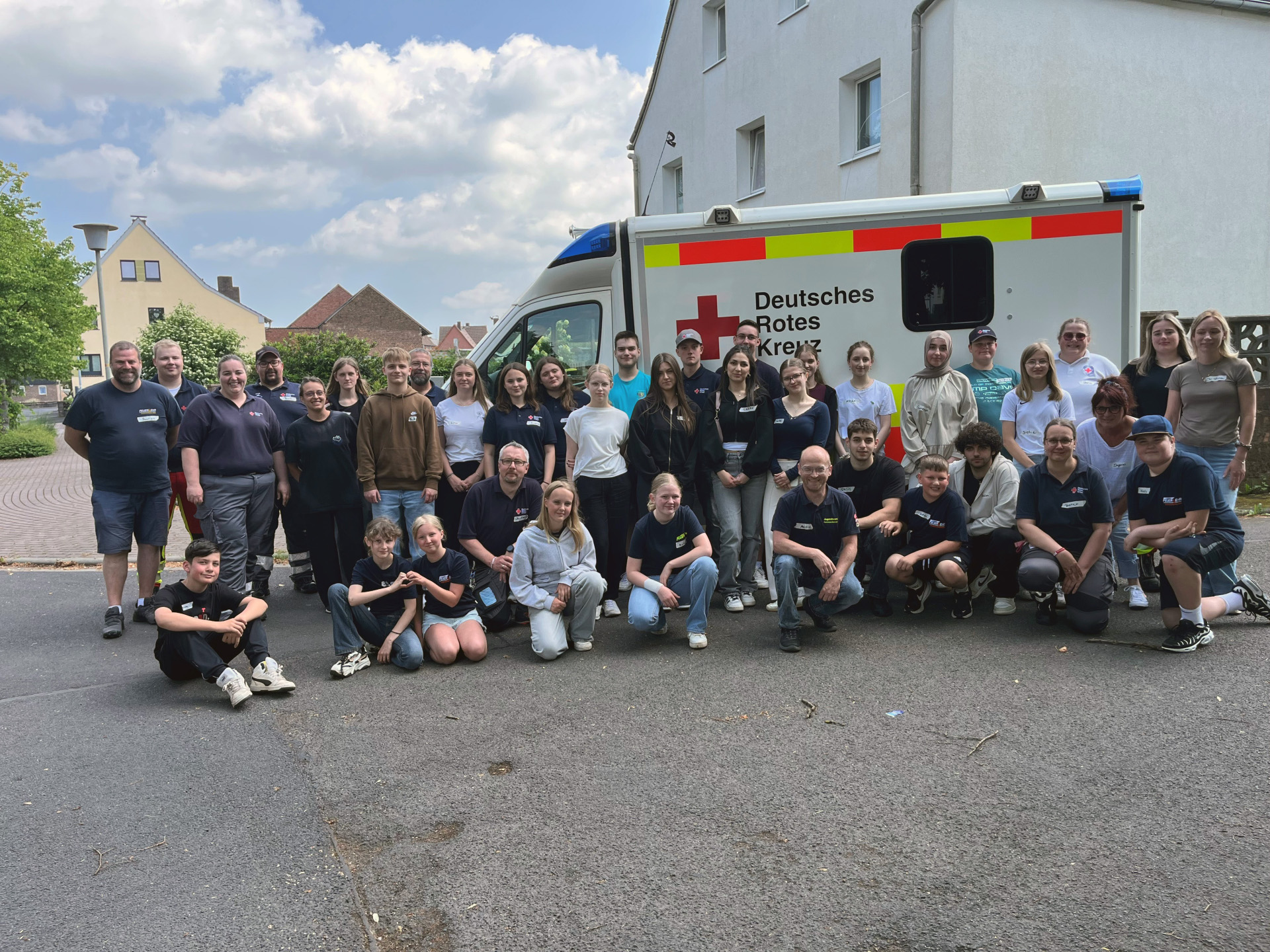Gruppenfoto vor einem Einsatzfahrzeug des Deutschen Roten Kreuzes: Rund 40 Jugendliche und Erwachsene stehen und knien gemeinsam auf einer Straße vor dem Fahrzeug und schauen in die Kamera. Die Gruppe besteht aus Teilnehmenden und Betreuenden eines Workshops von FSSJH, Feuerwehr und DRK. Im Hintergrund sind Wohnhäuser, Bäume und ein teilweise bewölkter Himmel zu sehen. Die Stimmung wirkt freundlich und gemeinschaftlich.