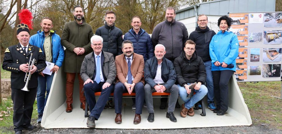 Eine Gruppe von elf Personen posiert im Freien für ein gemeinsames Foto. Sie stehen und sitzen in einer großen, umfunktionierten Baggerschaufel, die als Bank dient. Die Personen tragen überwiegend Alltags- und Outdoor-Kleidung; einige sitzen vorne, andere stehen dahinter. Links steht ein Mann in einer traditionellen Uniform mit schwarzer Jacke, Abzeichen und einem Hut mit rotem Federbusch und hält eine Trompete. Im Hintergrund sind kahle Bäume und eine Informationstafel mit Bildern und Texten zu sehen. Die Stimmung wirkt freundlich und offiziell, wie bei einer Einweihung oder einem lokalen Ereignis.