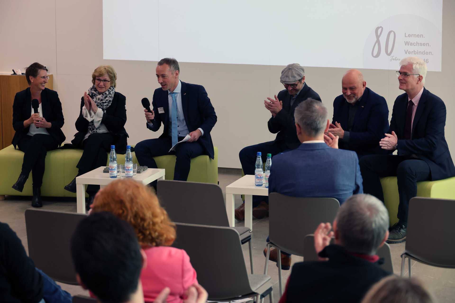 Auf dem Bild sind auf zwei grünen Sitzgelegenheiten insgesamt sechs Personen zu sehen. Auf einem kleinen Tisch vor ihnen stehen einige Wasserflaschen. Im Hintergrund ist eine Leinwand zu erkennen.