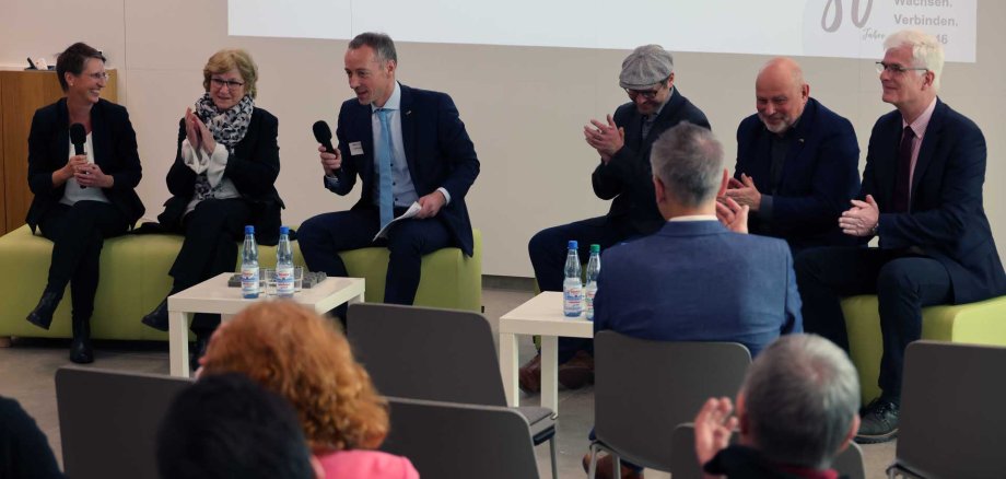 Auf dem Bild sind auf zwei grünen Sitzgelegenheiten insgesamt sechs Personen zu sehen. Auf einem kleinen Tisch vor ihnen stehen einige Wasserflaschen. Im Hintergrund ist eine Leinwand zu erkennen.