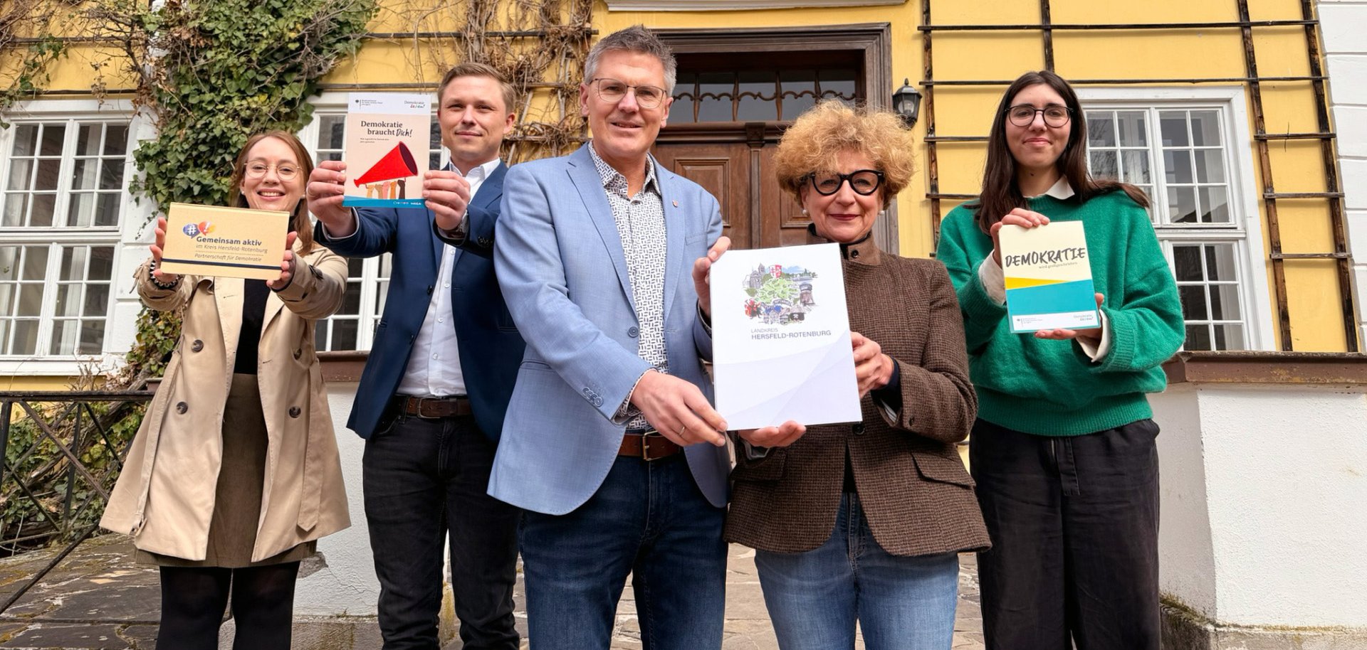 Fünf Personen stehen vor einem gelb gestrichenen Gebäude mit weißen Fenstern und halten Broschüren und Materialien zum Thema Demokratie in die Kamera. In der Mitte präsentieren zwei Personen eine Mappe mit einem Wappen, während die anderen links und rechts daneben Informationshefte hochhalten. Im Hintergrund sind Kletterpflanzen an der Fassade zu sehen.