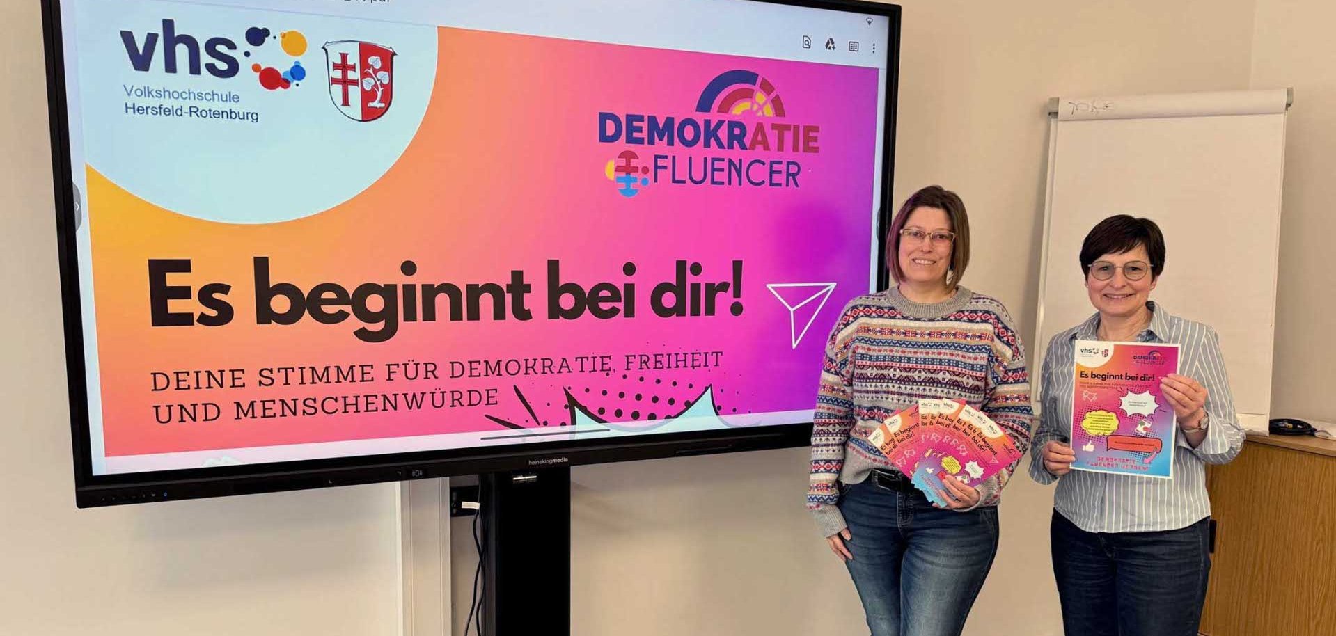 Zwei Frauen stehen in einem Seminarraum neben einem großen Bildschirm. Auf dem Bildschirm ist eine Präsentationsfolie der Volkshochschule Hersfeld-Rotenburg mit dem Titel „Demokratie-Fluencer“ und dem Slogan „Es beginnt bei dir! Deine Stimme für Demokratie, Freiheit und Menschenwürde“. Beide Frauen halten bunte Flyer der Kampagne in den Händen. Neben ihnen steht ein Flipchart.