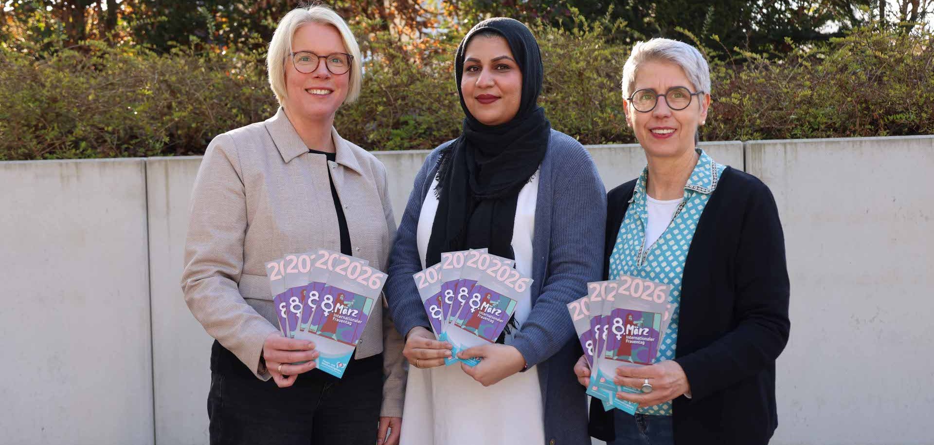 Drei Frauen stehen vor einer Hecke. Die Sonne scheint. Sie halten alle mehrere Flyer zum Internationalen Frauentag 2026 in der Hand.