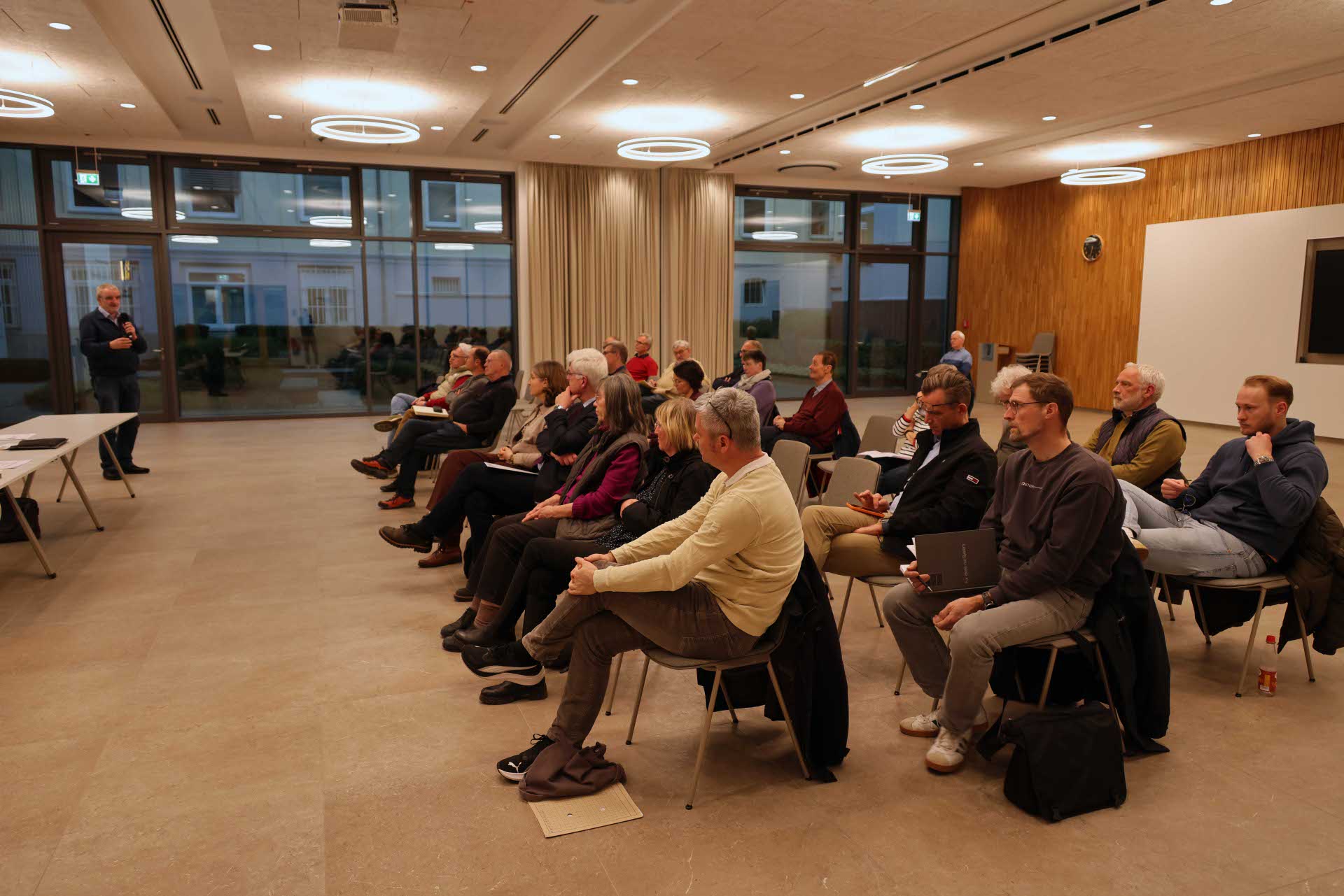 Menschen sitzen in einem Sitzungssaal in Reihen angeordnet. Vorne steht eine Person mit Mikrofon.
